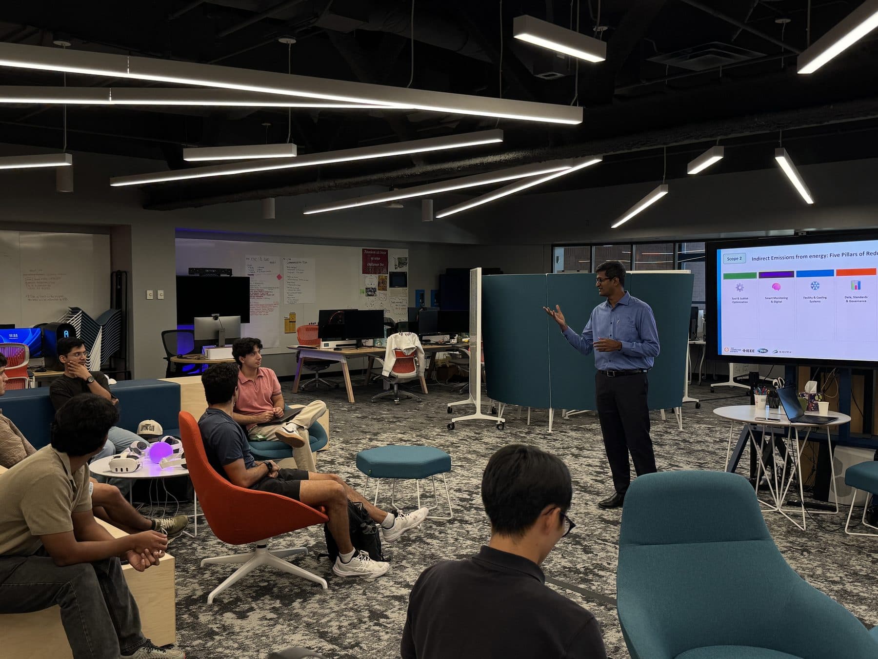 A speaker presents next to a large screen while students listen from chairs and benches in an open, modern room.