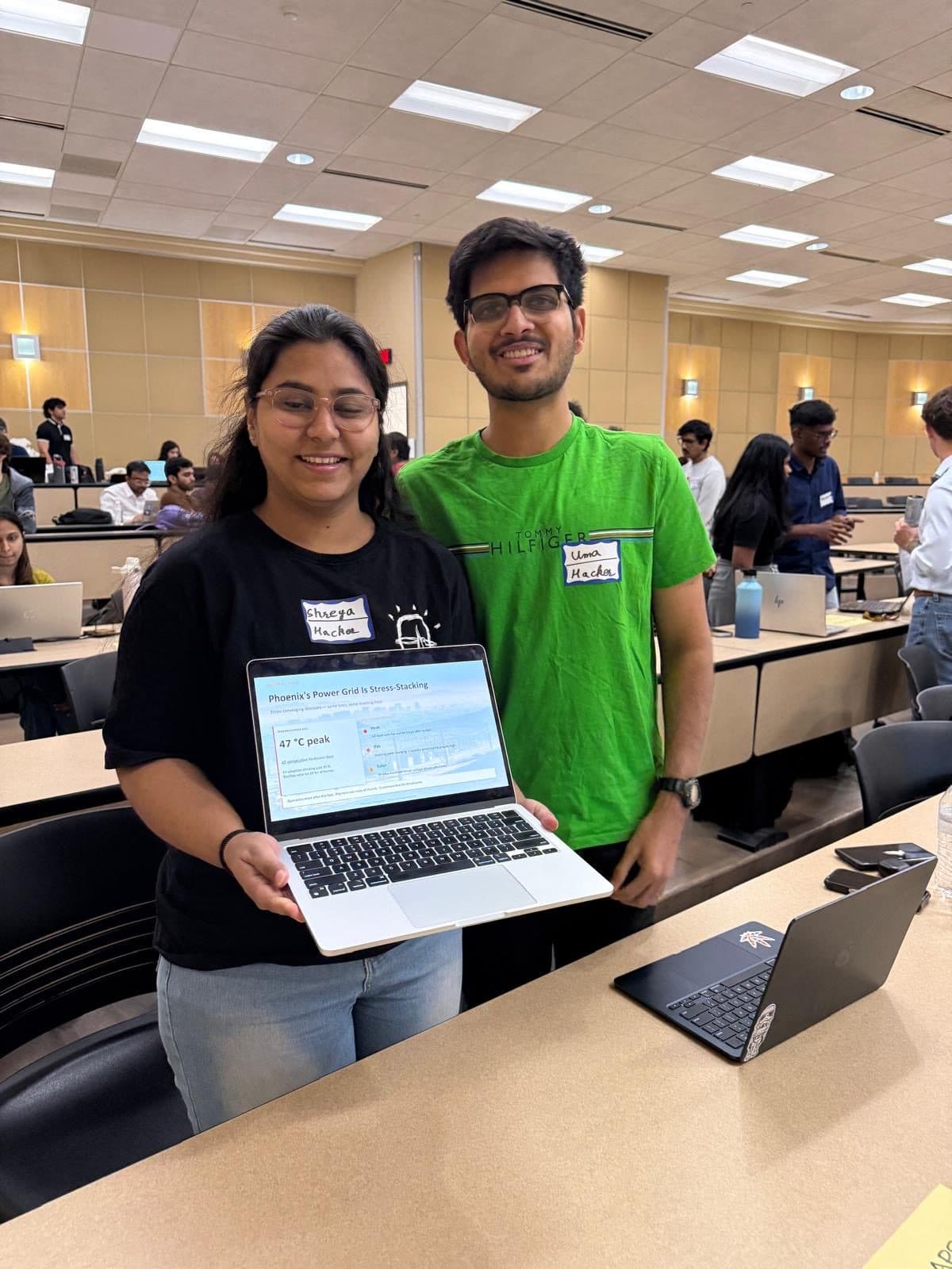 Two students hold up a laptop showing their project dashboard at their team's table.