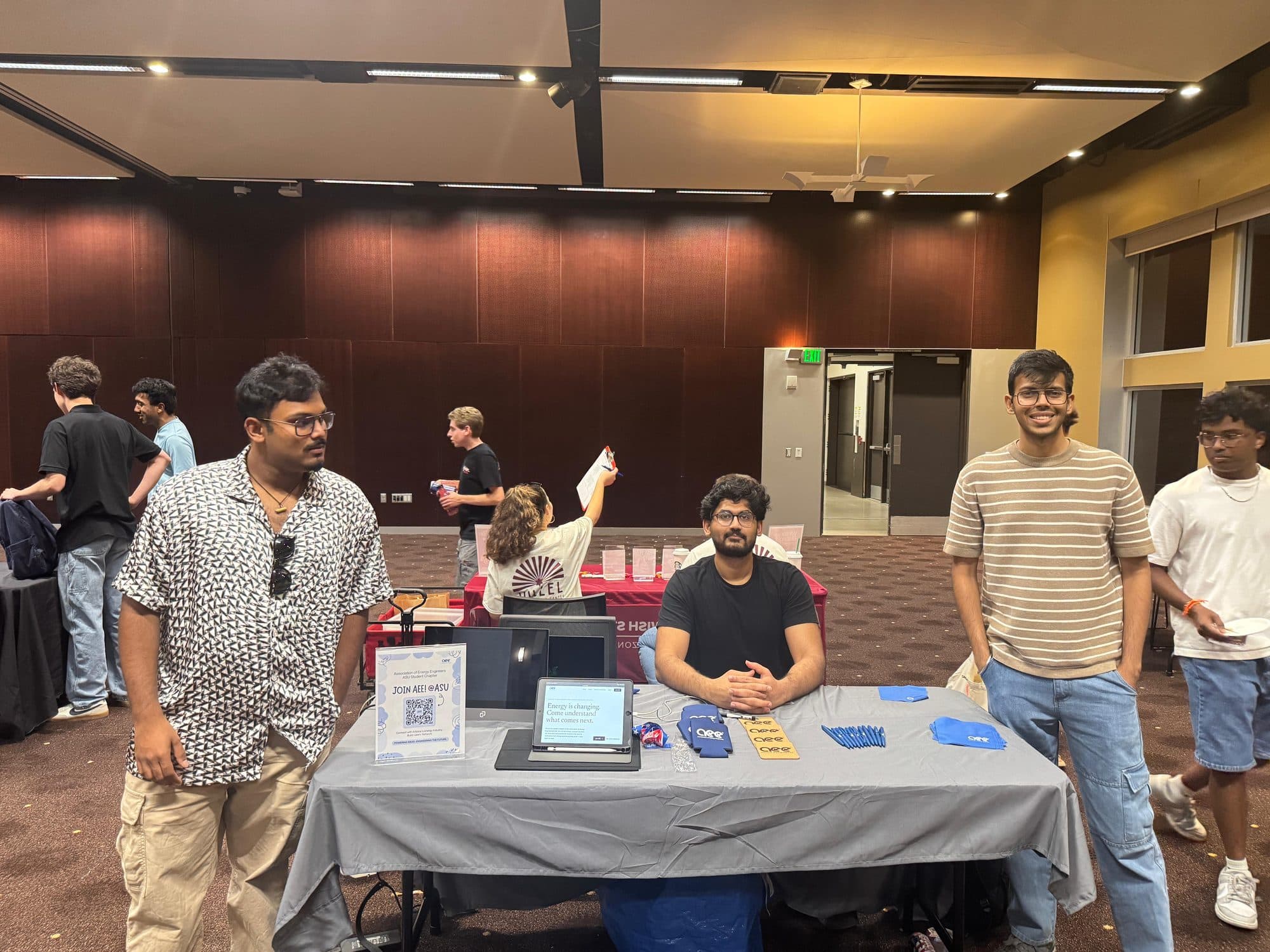 Three chapter officers at their table during a student organization fair, two standing and one seated, with flyers, pens and giveaways laid out and a tablet showing the chapter website.