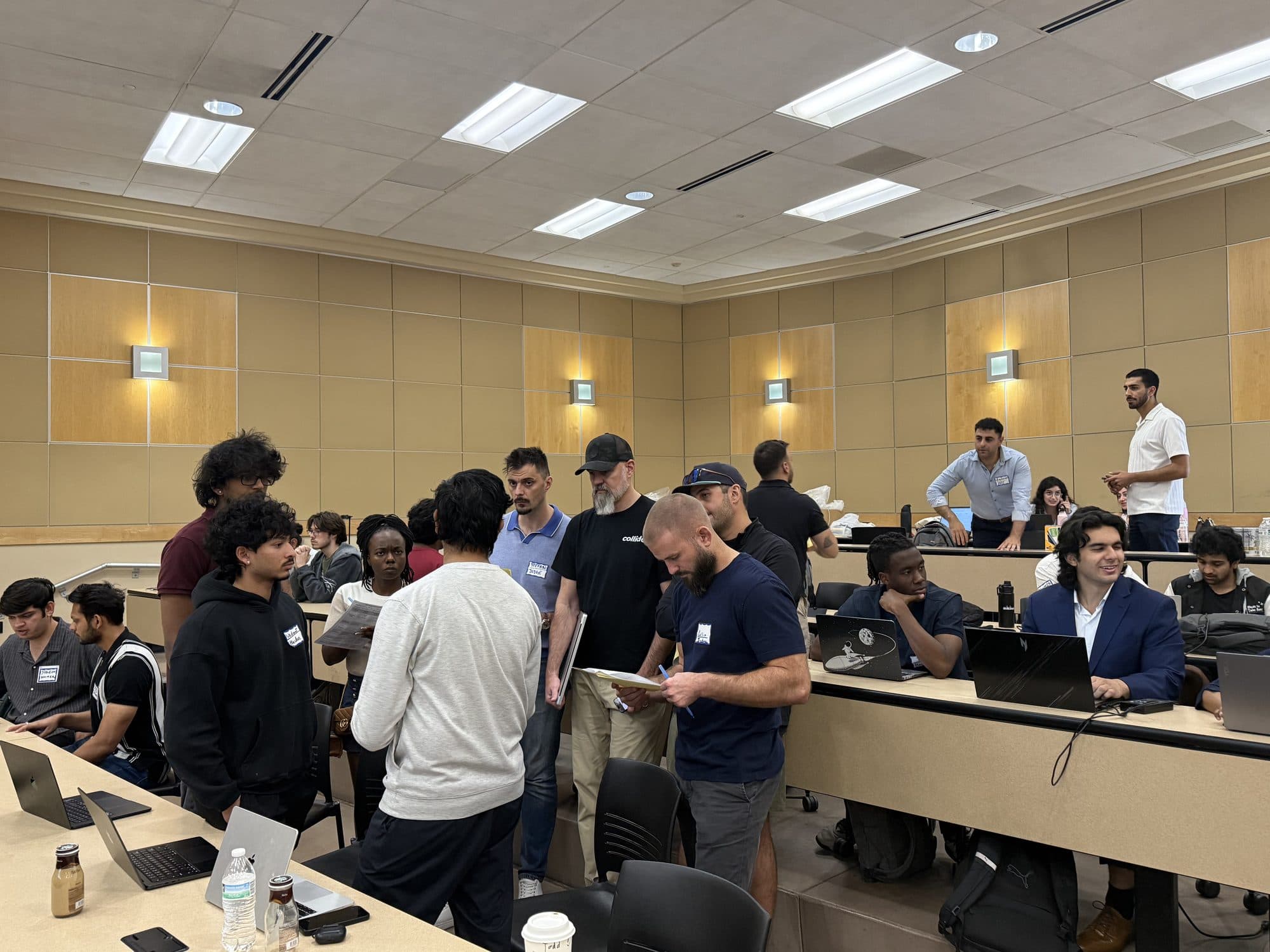 A mentor leans over a bench to review a team's work on a laptop while other participants continue working nearby.