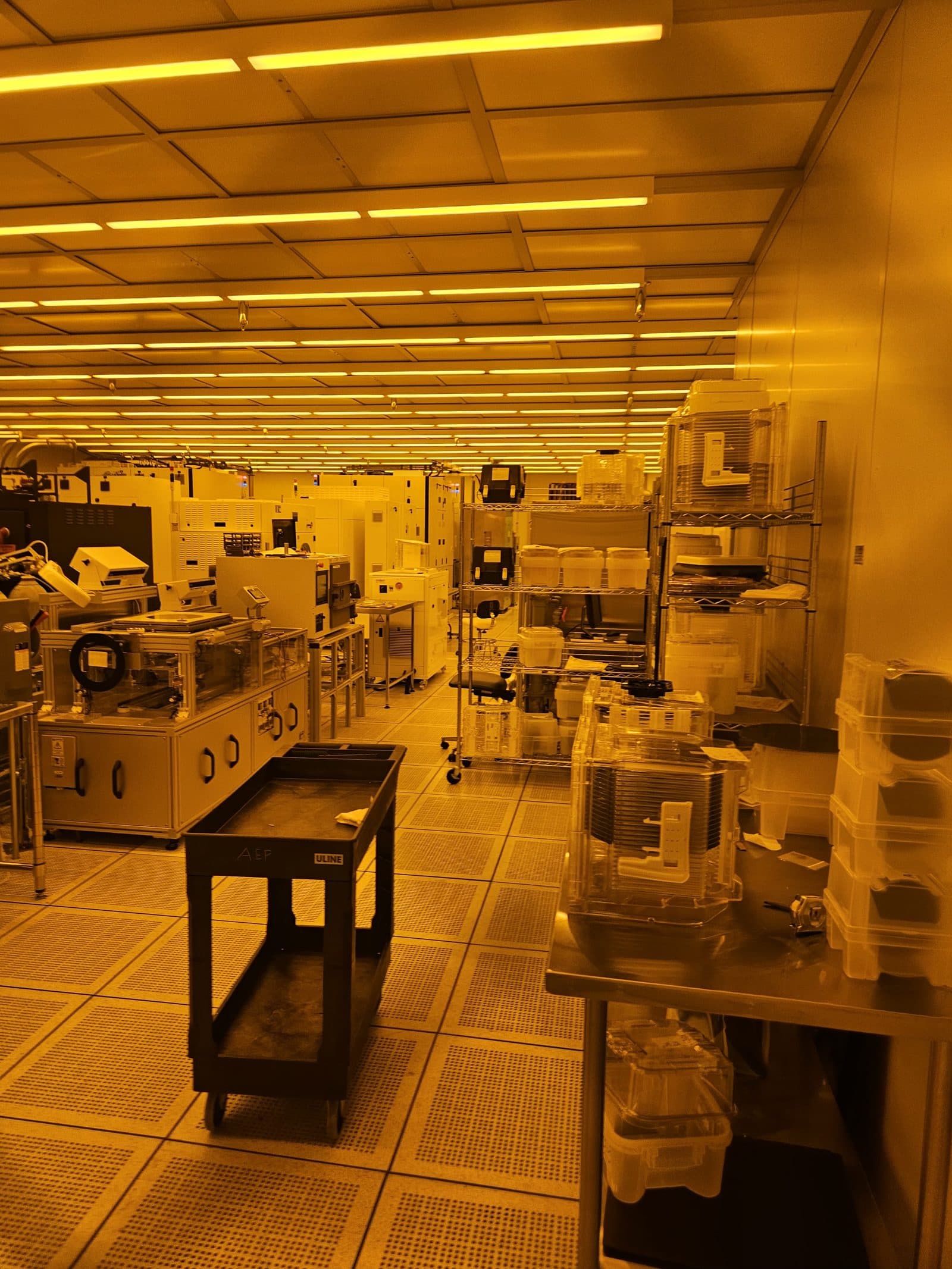 A long cleanroom bay under amber lighting, lined with process equipment, shelving and a cart.