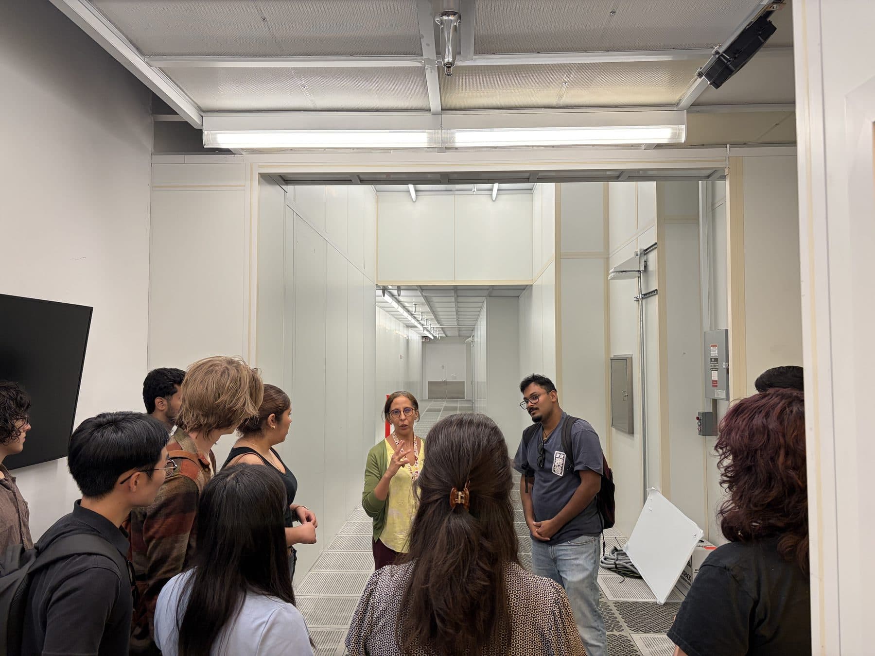 Students stand in a white corridor outside the cleanroom, gathered around two people who are talking with them.